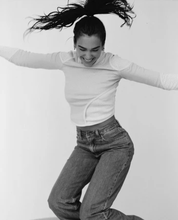 A woman jumps in a black-and-white studio shot wearing jeans and a long-sleeved shirt, smiling with her hair in a high ponytail.