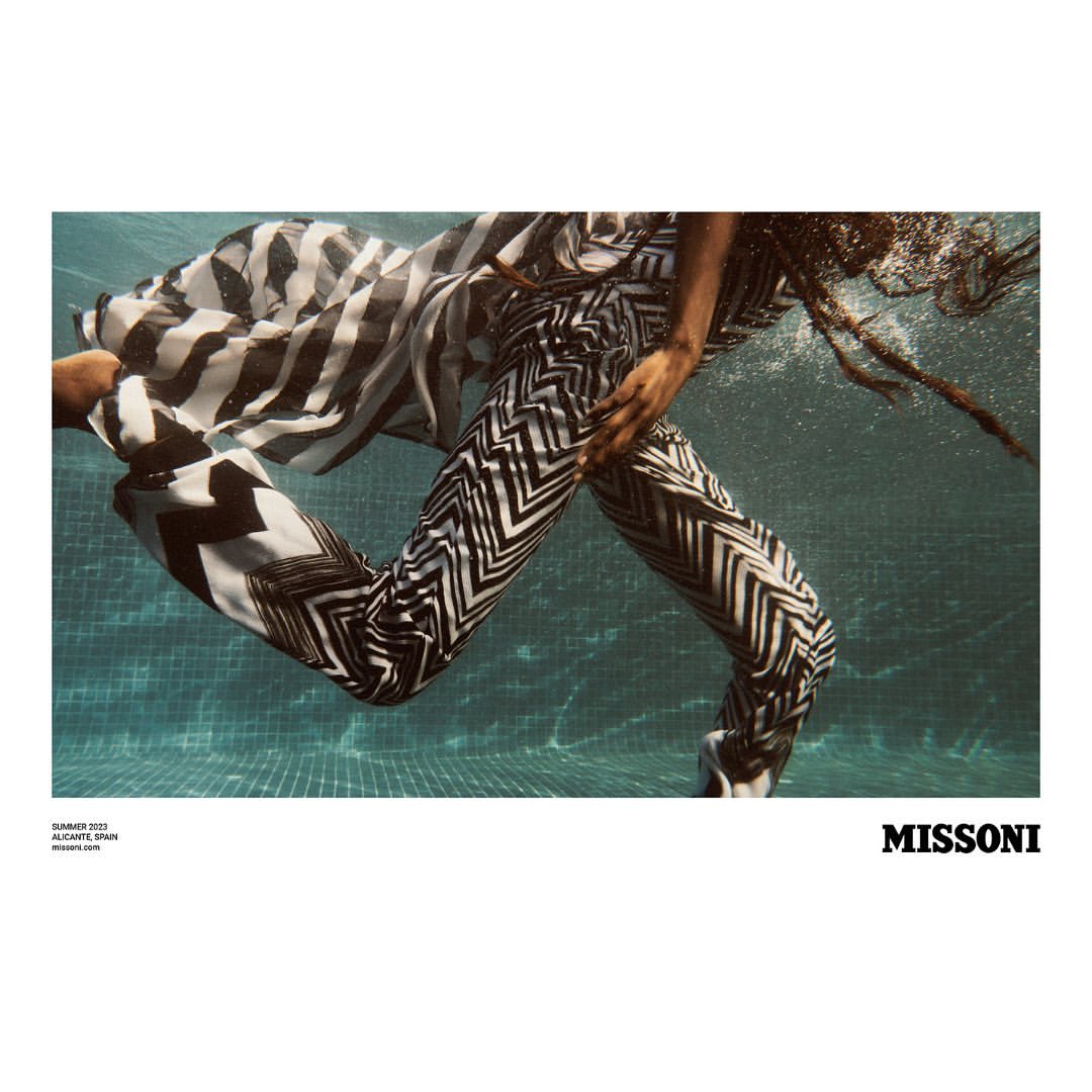 Woman in patterned pants swims underwater in a pool. Fashion shot with summer 2022 branding of Alicante, Spain, and the Missoni logo.
