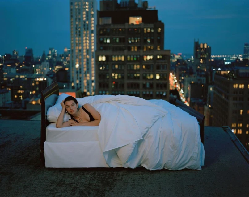Woman in a black top and jewelry lies on a bed in a white comforter on a rooftop in a city at night with lights.