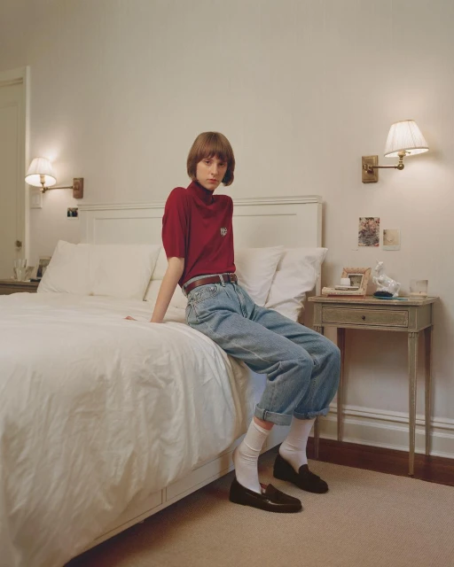 Young adult poses on a white bed in a bedroom, wearing a maroon shirt, jeans, white socks, and loafers near a wooden nightstand.