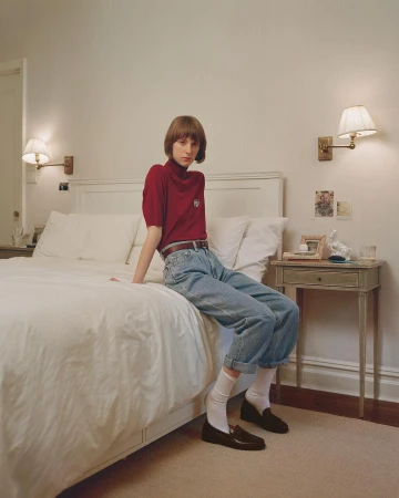 Young adult poses on a white bed in a bedroom, wearing a maroon shirt, jeans, white socks, and loafers near a wooden nightstand.
