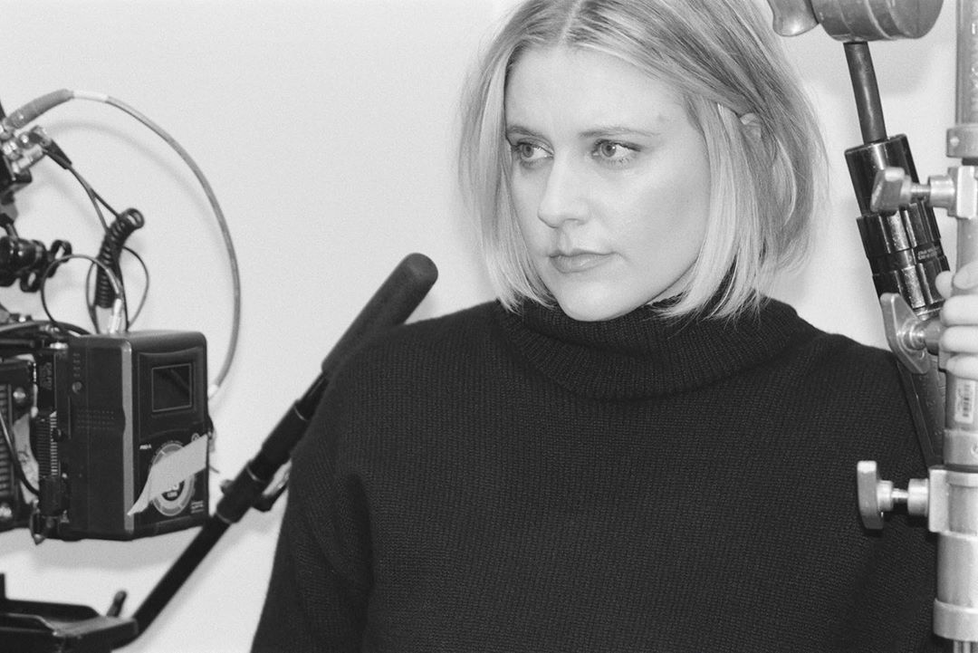 Monochrome portrait of a blonde woman in a black turtleneck next to camera equipment in a bright indoor photo studio setting.