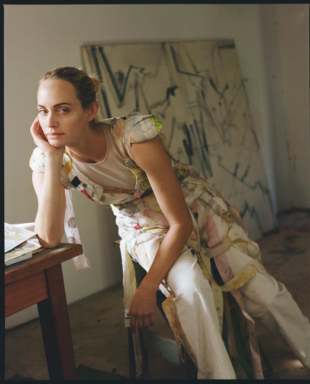 Blonde woman in patterned dress poses beside table and abstract painting in art studio with soft lighting and neutral colors.