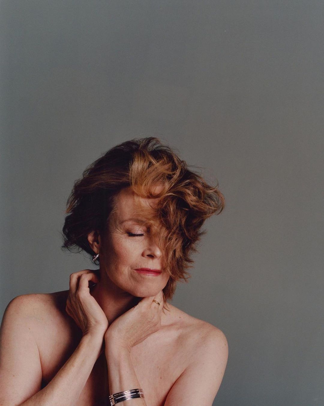 Studio portrait of a middle-aged woman with bare shoulders against a grey background. She has short auburn hair and is wearing jewelry.