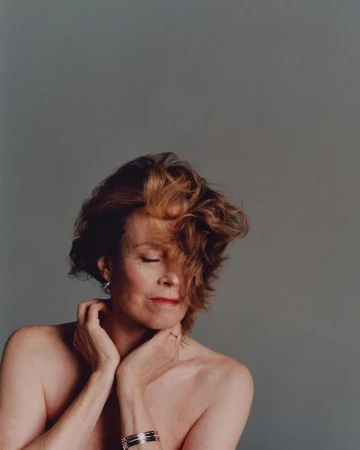 Studio portrait of a middle-aged woman with bare shoulders against a grey background. She has short auburn hair and is wearing jewelry.