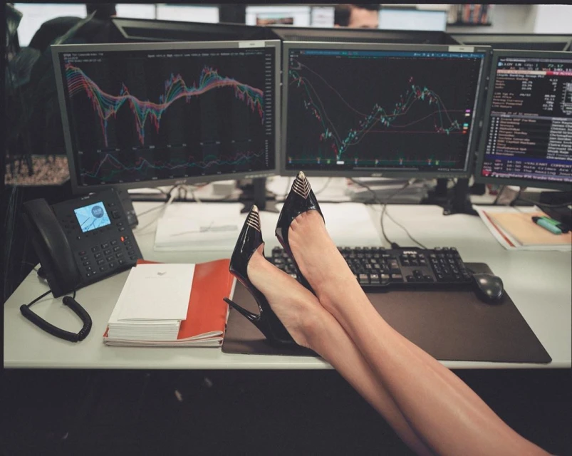 Woman's legs up on a desk in an office with multiple computer screens displaying trading charts, a black telephone, and a keyboard.