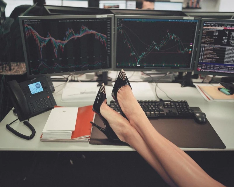 Office scene featuring a woman's legs resting on a desk with financial charts on computer monitors, a phone, and office supplies.