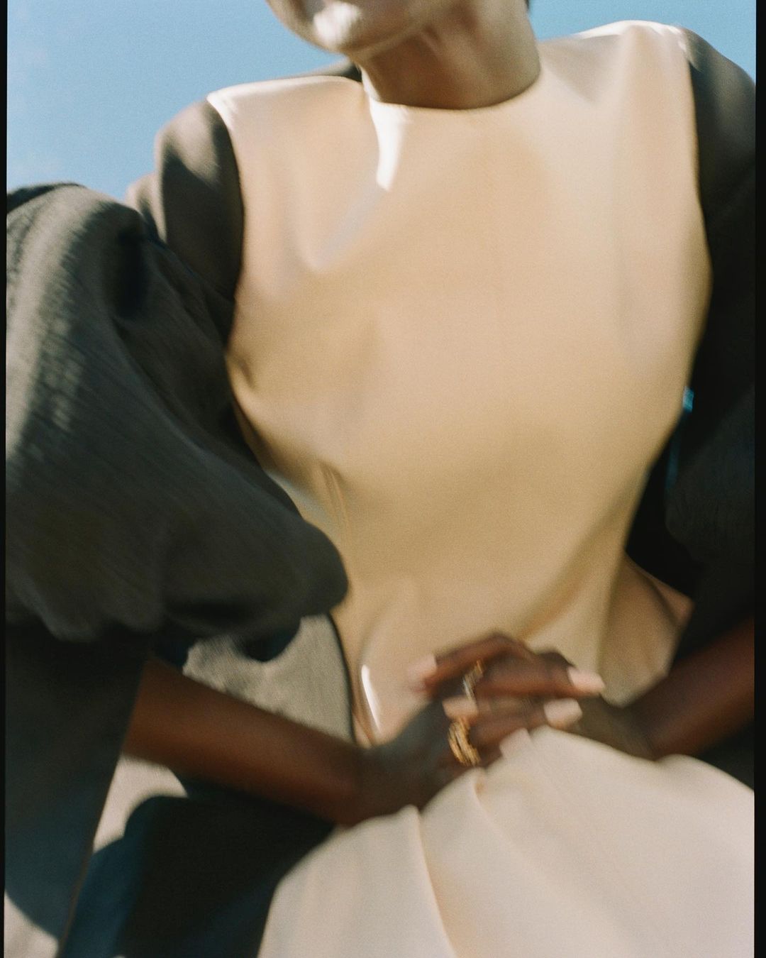 Elegant Black woman in a light tan and black dress sitting in the sun with crossed hands, wearing gold rings, against a blue sky.