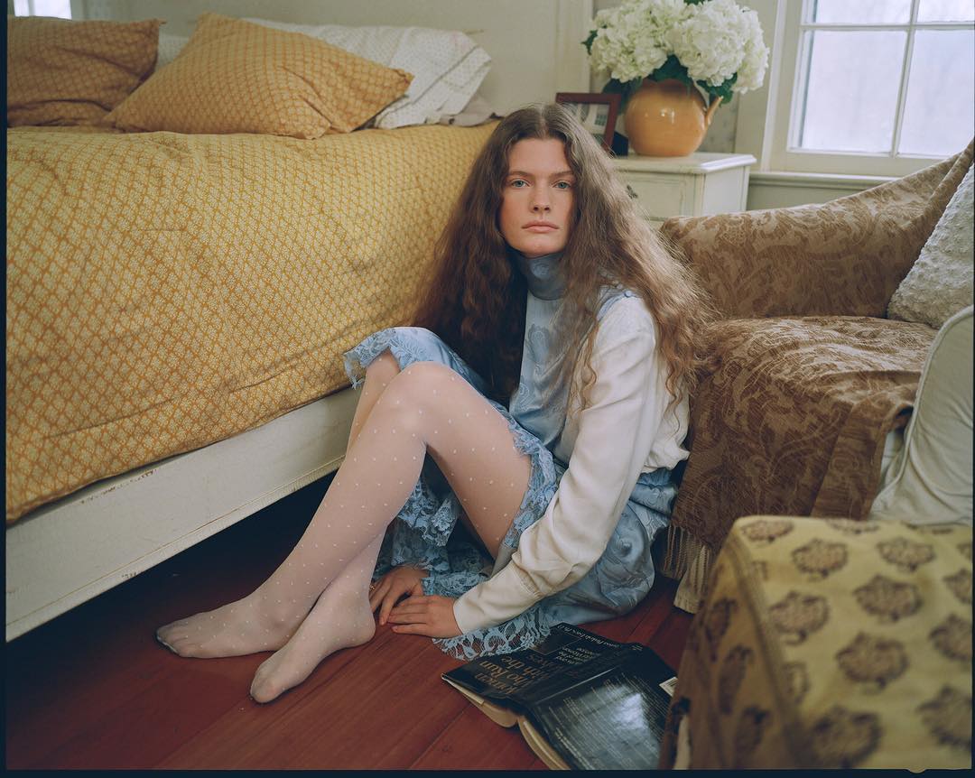 Woman with long wavy red hair sits on the floor in a blue and white dress with sheer white dotted tights in a natural light setting.