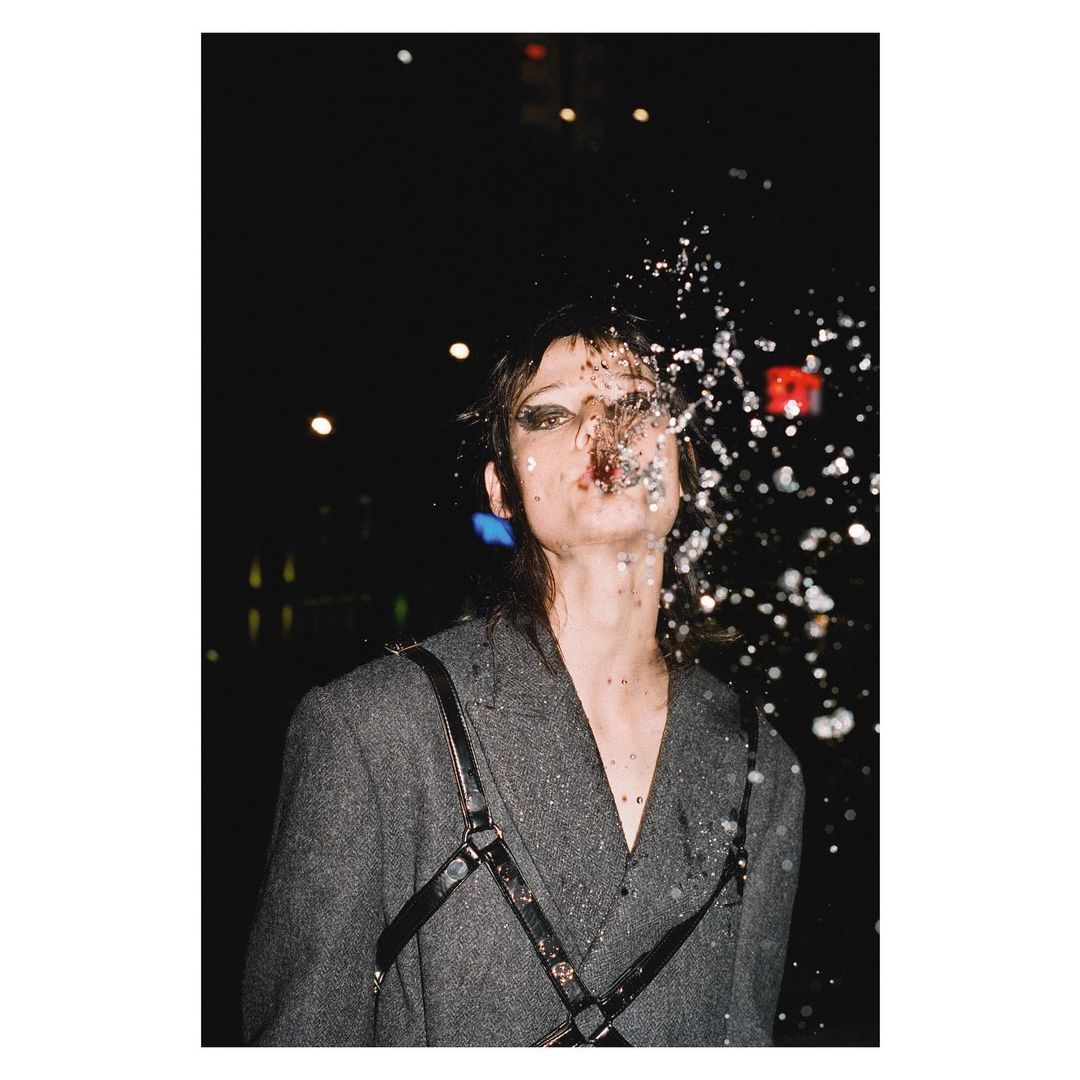 Woman in a grey blazer stands outside with water splashing on her face under dark lighting with lights, creating a dramatic effect.
