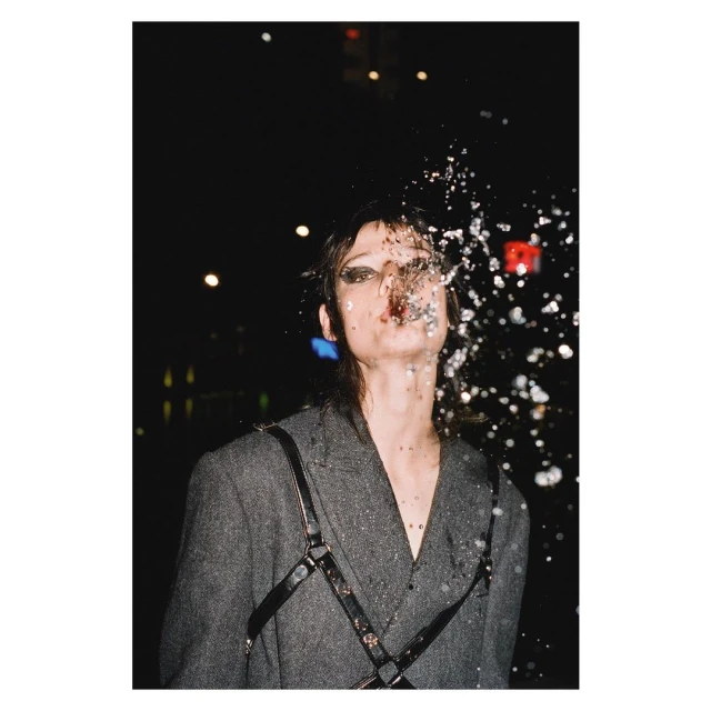 Woman in a grey blazer stands outside with water splashing on her face under dark lighting with lights, creating a dramatic effect.