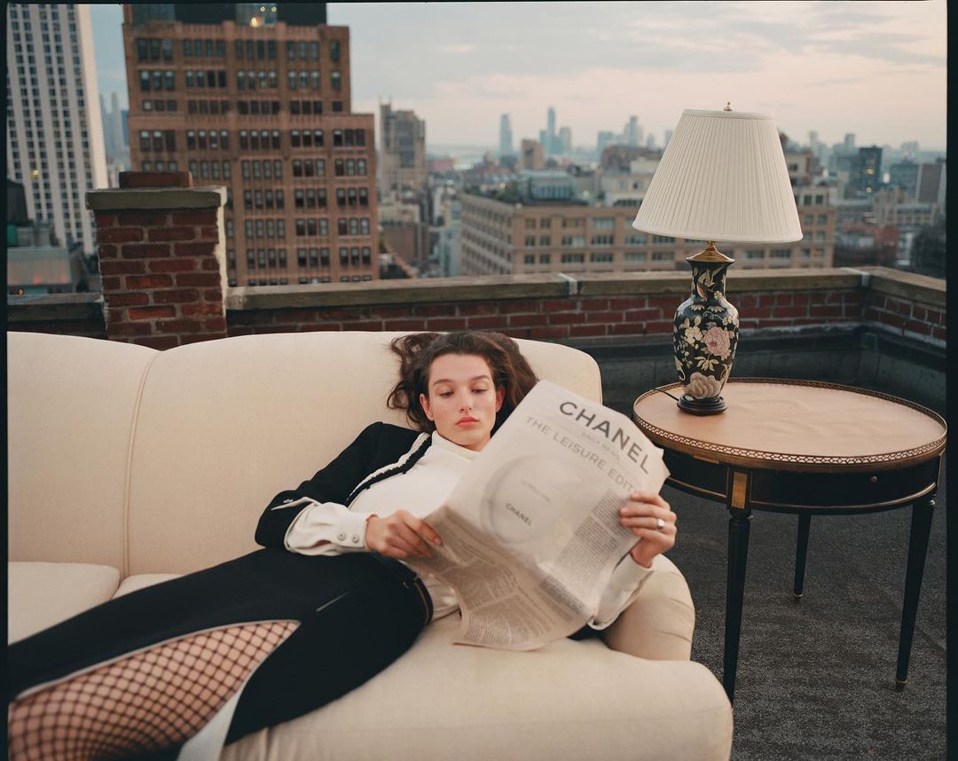 A woman lounges on a cream sofa on a rooftop in an urban setting, casually reading a newspaper next to a table lamp and round accent table.