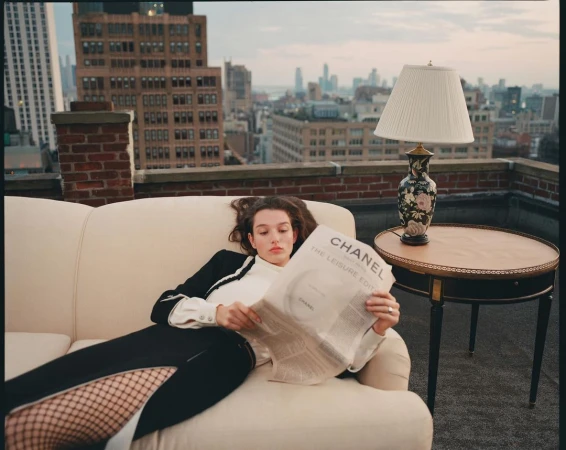 A woman lounges on a cream sofa on a rooftop in an urban setting, casually reading a newspaper next to a table lamp and round accent table.