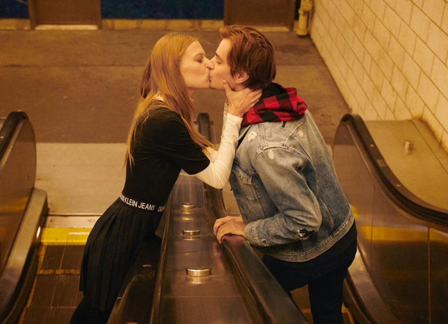 A couple shares a kiss on an escalator. The woman wears a black dress, and the man is in a denim jacket, with yellow tiled walls.