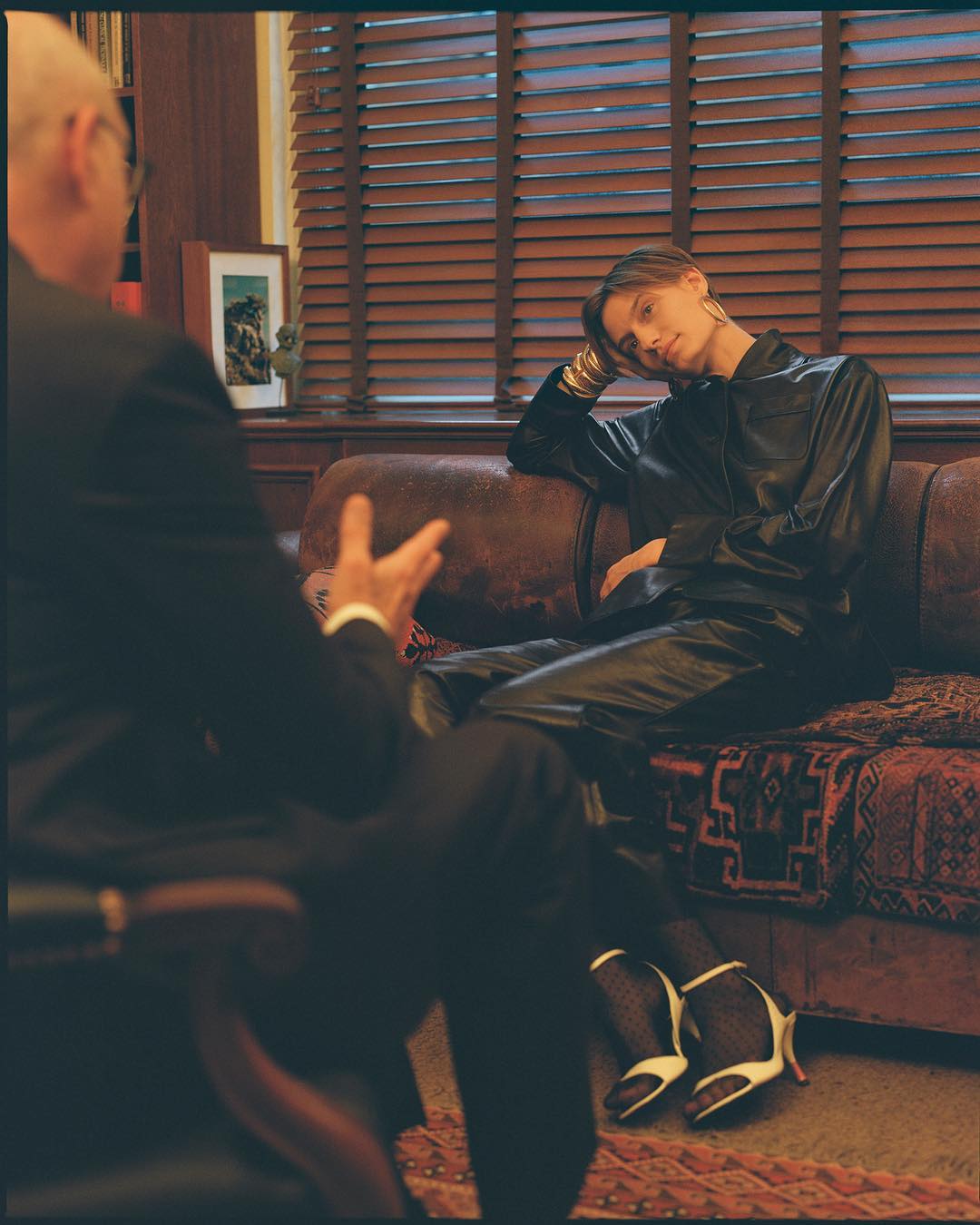 Woman in leather outfit poses on a brown leather couch, consulting with a man in a suit in an indoor setting with blinds.