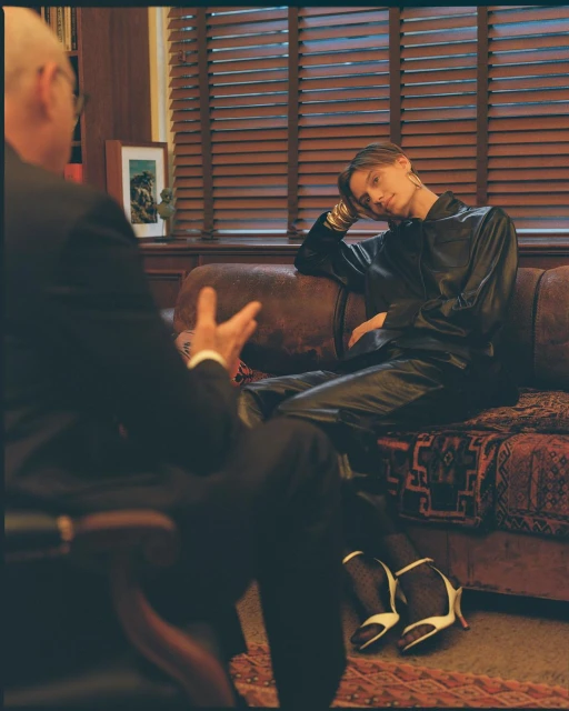 Woman in leather outfit poses on a brown leather couch, consulting with a man in a suit in an indoor setting with blinds.