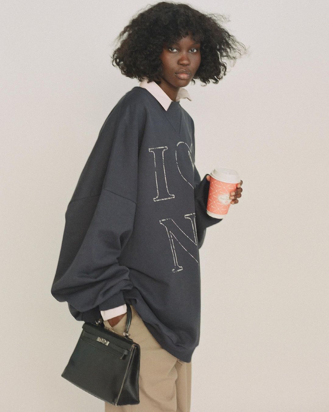 A woman with dark curly hair holds a coffee cup and black purse, wearing an oversized navy sweatshirt and khaki pants against a white background.