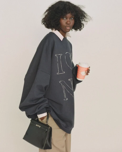 A woman with dark curly hair holds a coffee cup and black purse, wearing an oversized navy sweatshirt and khaki pants against a white background.