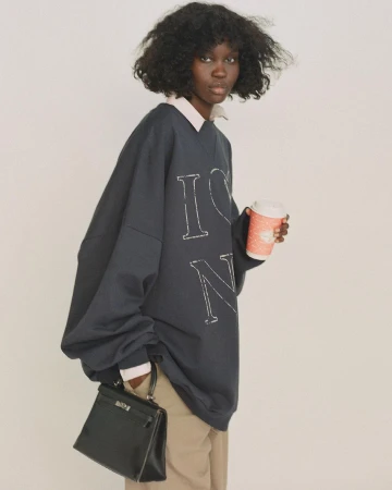 A woman with dark curly hair holds a coffee cup and black purse, wearing an oversized navy sweatshirt and khaki pants against a white background.
