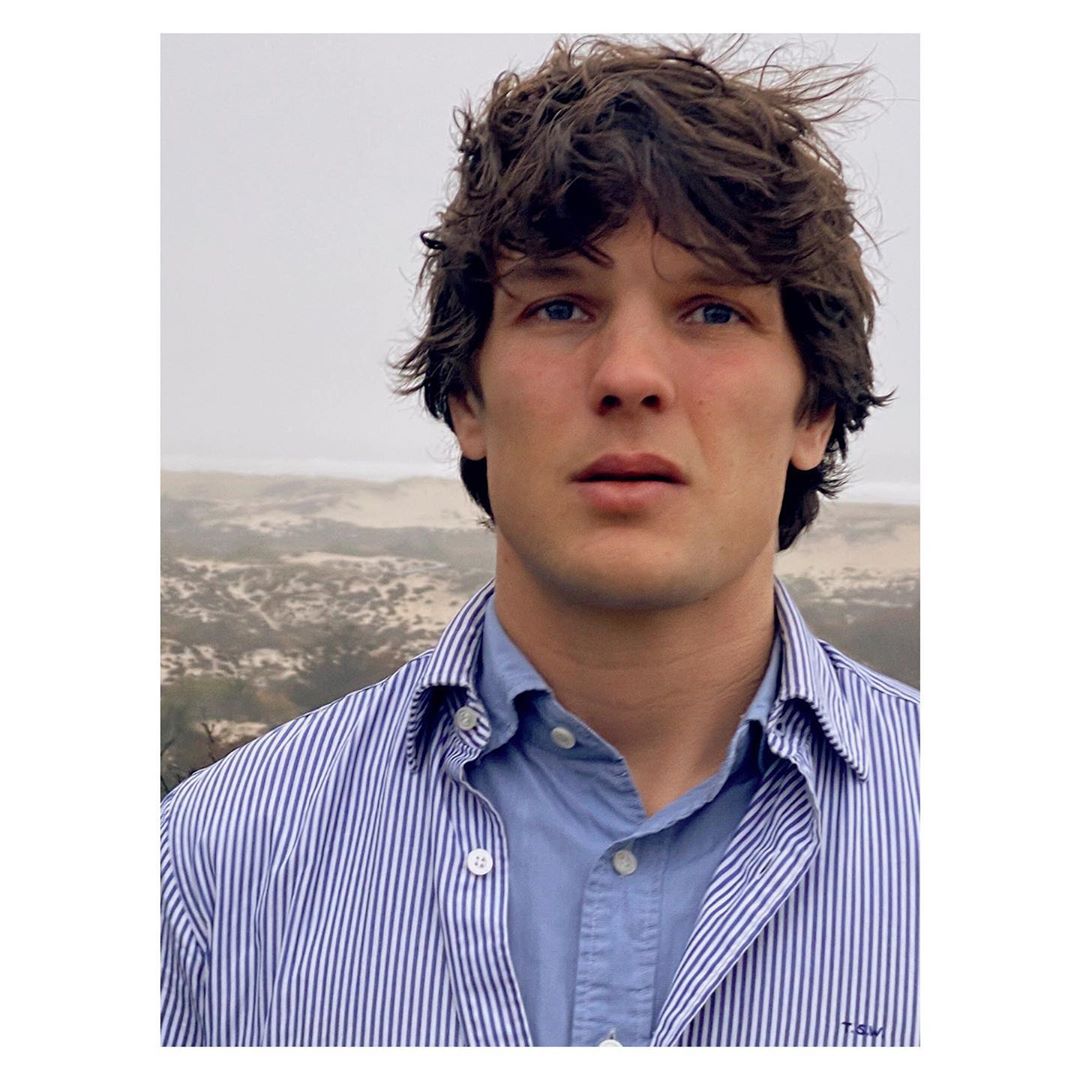 Portrait of a light-skinned adult male with dark, wavy hair, wearing a blue collared shirt, set against an outdoor background.