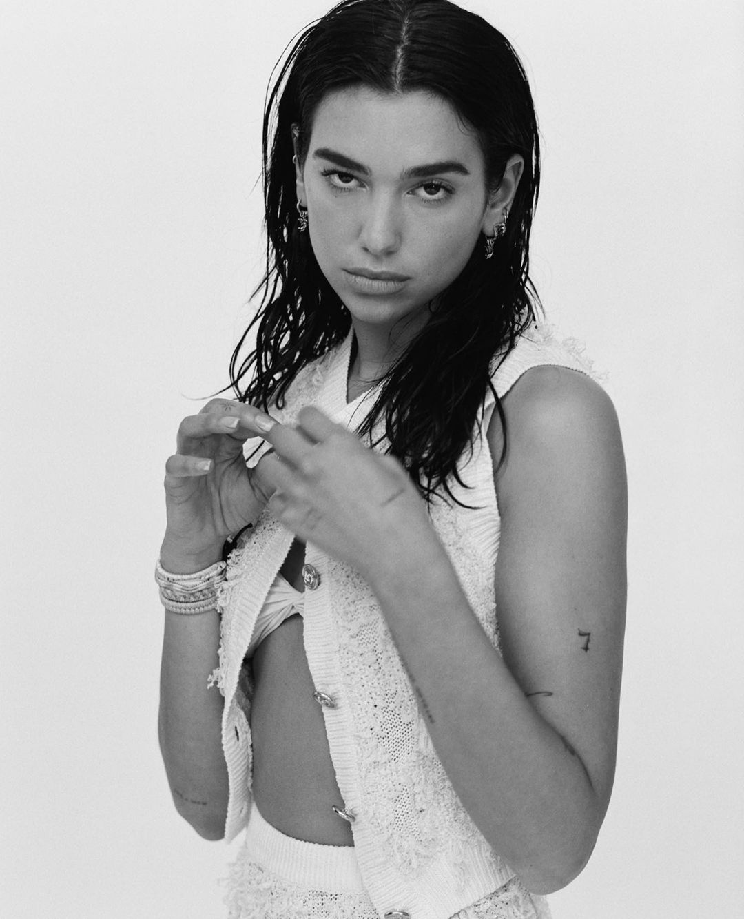 Monochrome portrait of Dua Lipa in a white tweed outfit against a white background, showcasing her jewelry and arm tattoos.
