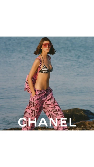 A woman in a bikini top and patterned pants walks along a sunny beach with a pink backpack, with blue ocean in the background.