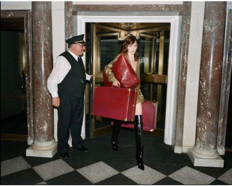 A fashionable model with red luggage emerges from revolving doors, aided by a doorman in uniform; marble columns frame the scene.