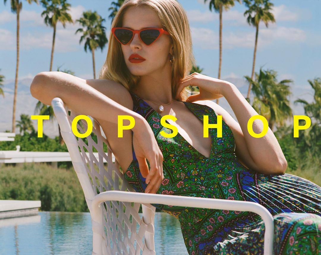 A blonde model wearing heart-shaped sunglasses and a floral dress sits on a white chair in front of a pool and palm trees.