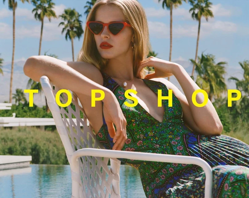 A blonde model wearing heart-shaped sunglasses and a floral dress sits on a white chair in front of a pool and palm trees.