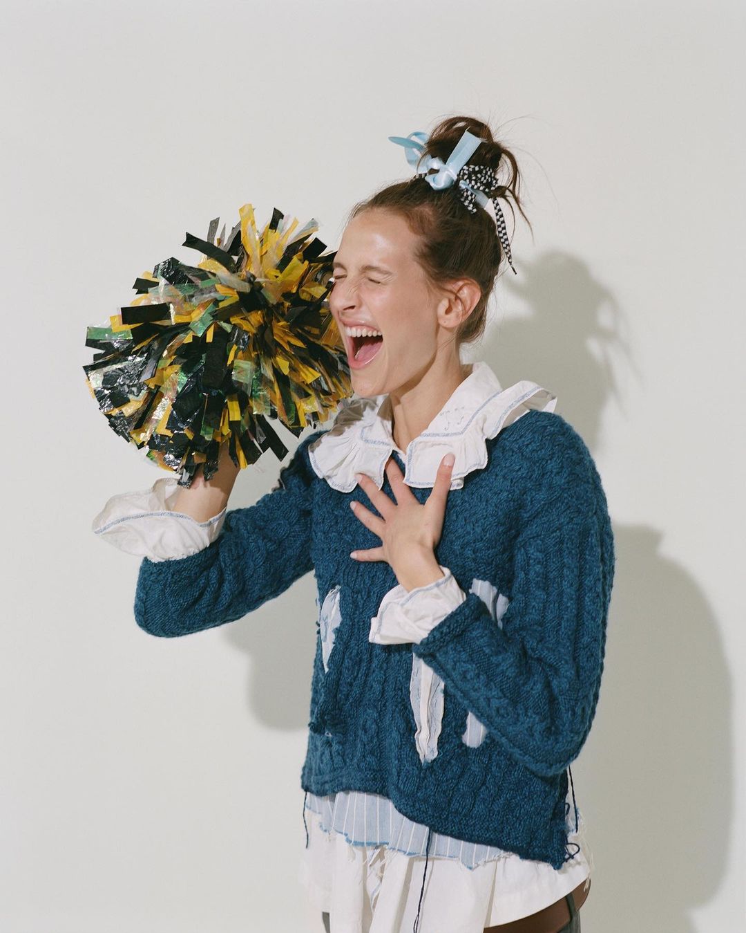 Enthusiastic young woman in a blue cable knit sweater cheers with black and yellow pom-poms against a light neutral backdrop.