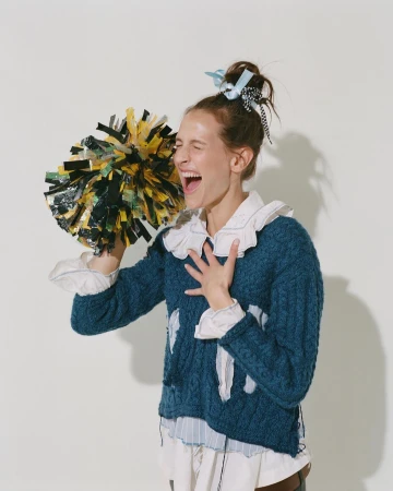 Enthusiastic young woman in a blue cable knit sweater cheers with black and yellow pom-poms against a light neutral backdrop.