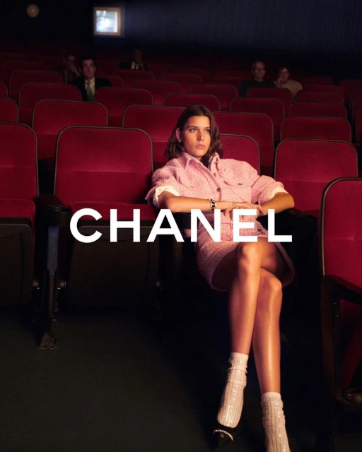 A solo woman in a pink tweed skirt suit sitting in a red-seated theatre, with logo-type text 'CHANEL' overlaid on the image.