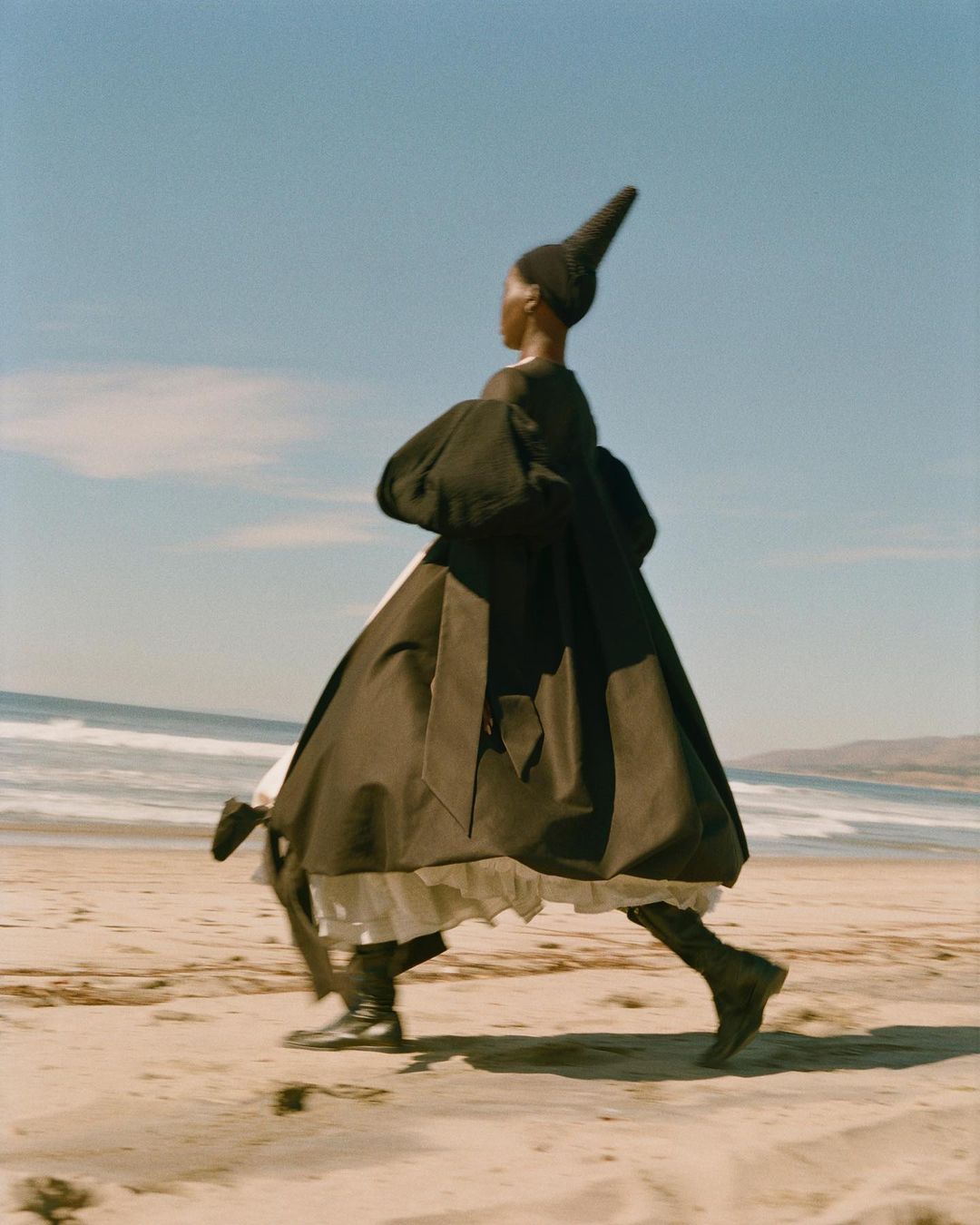 Fashion shot of a Black woman walking on a beach. She is wearing a dark cape-like dress with a tall, pointed hat, boots, with a sunny, ocean backdrop.