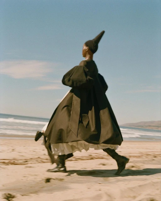 Fashion shot of a Black woman walking on a beach. She is wearing a dark cape-like dress with a tall, pointed hat, boots, with a sunny, ocean backdrop.