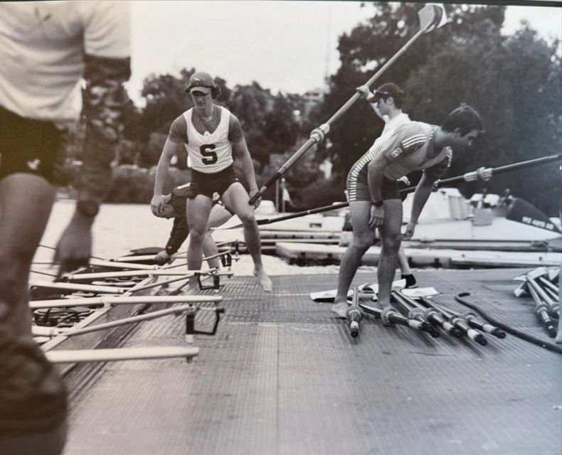 Rowing team members standing on a dock with oars in a black and white photograph. People are preparing to row on the water.