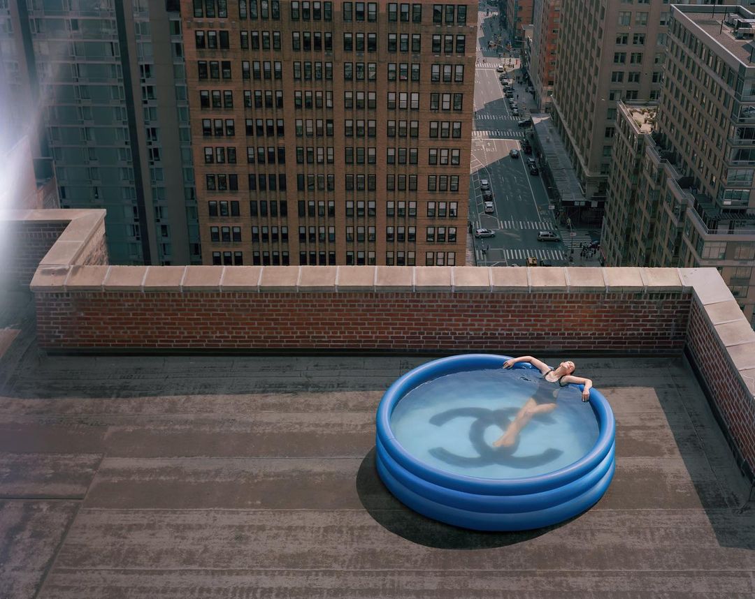 Woman in a blue kiddie pool with a Chanel logo, atop a rooftop in an urban setting. Tall buildings are visible in the background.