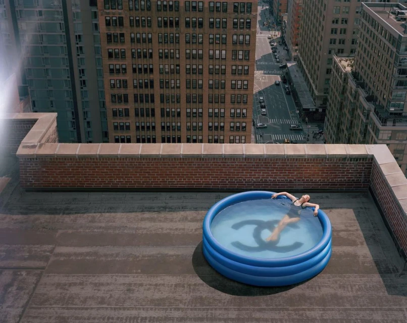 Woman in a blue kiddie pool with a Chanel logo, atop a rooftop in an urban setting. Tall buildings are visible in the background.