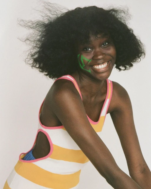 Smiling woman with dark skin and curly hair poses in a white and yellow striped dress with pink trim against a white background.