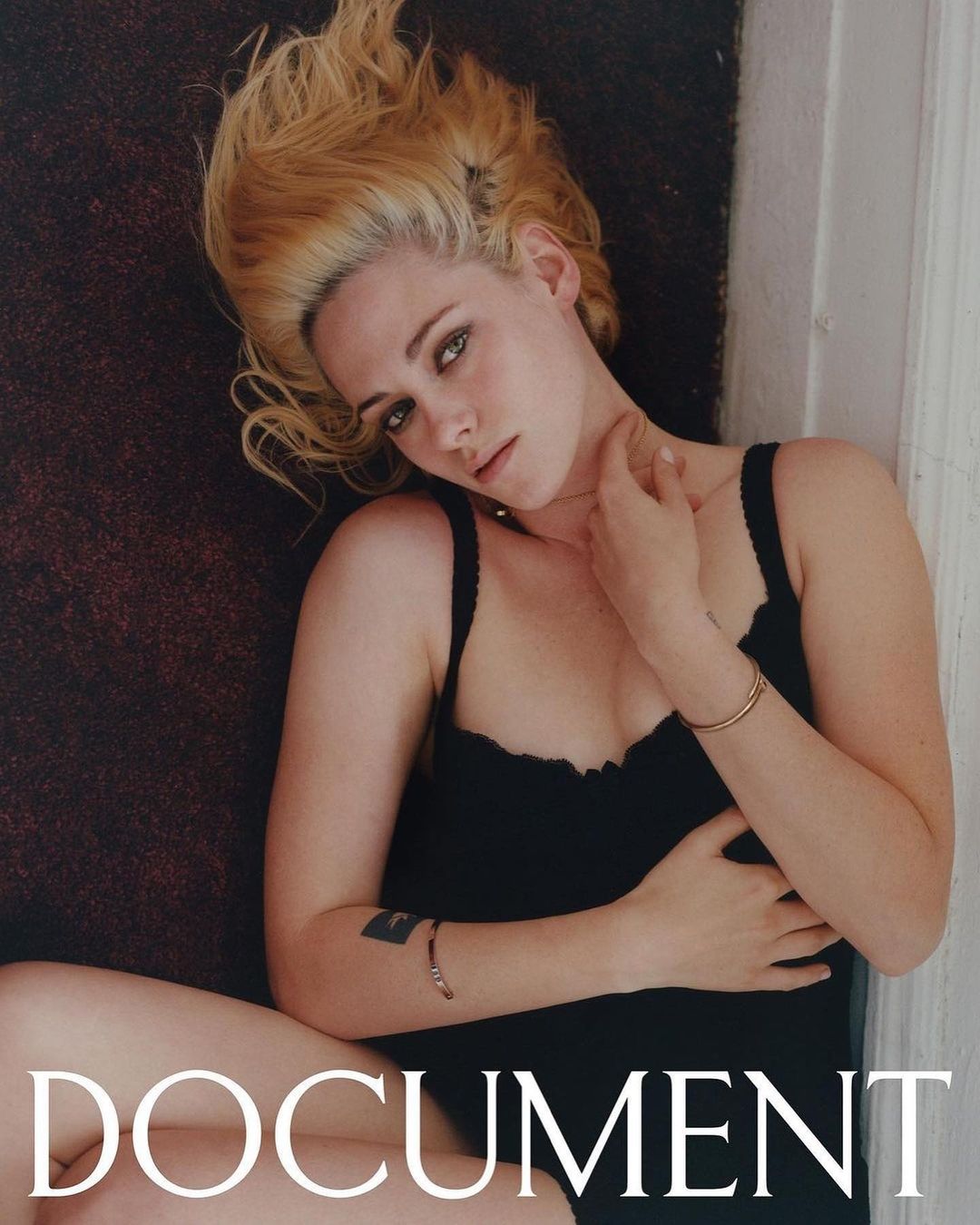 Kristen Stewart lying down wearing a black tank top with blonde hair spread out against a dark carpet and white wall