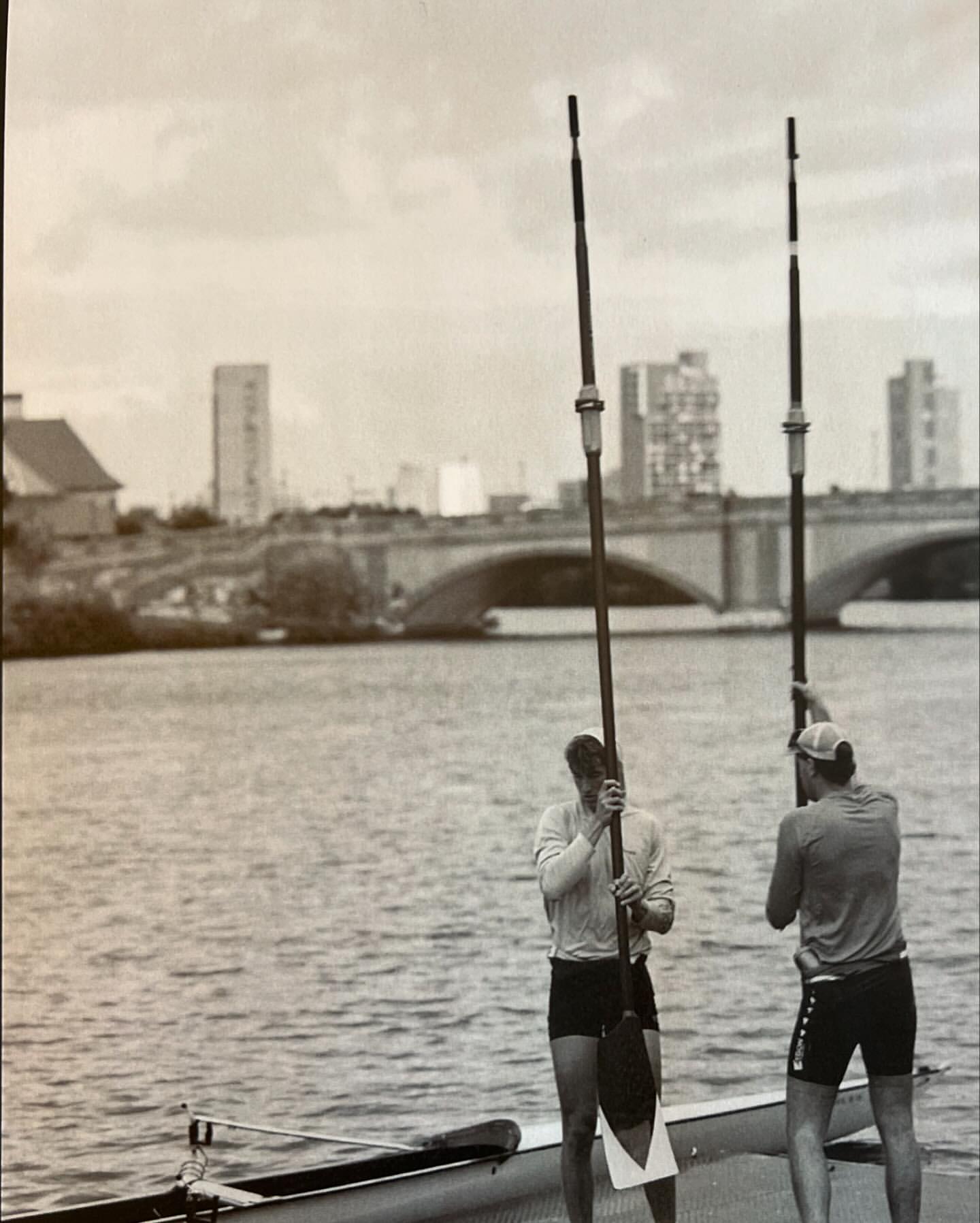 Two rowers stand with oars in a grayscale shot near a river, with a bridge and buildings in the background on an overcast day.