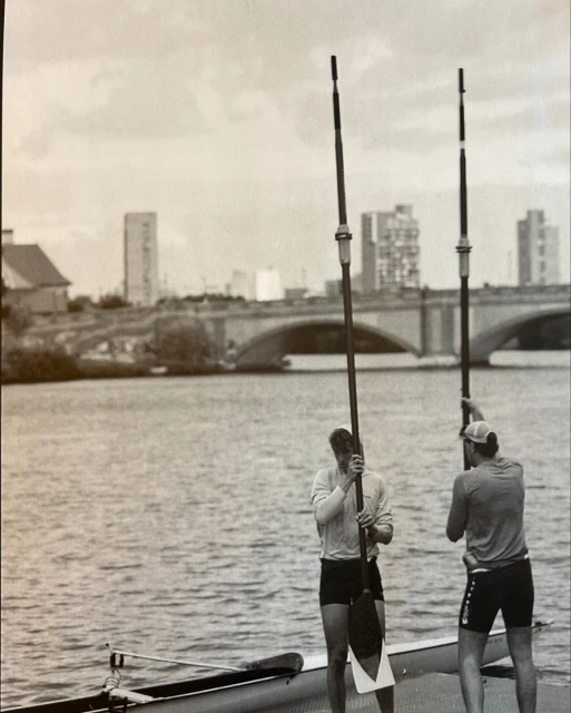 Two rowers stand with oars in a grayscale shot near a river, with a bridge and buildings in the background on an overcast day.