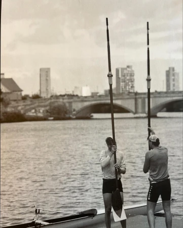 Two rowers stand with oars in a grayscale shot near a river, with a bridge and buildings in the background on an overcast day.