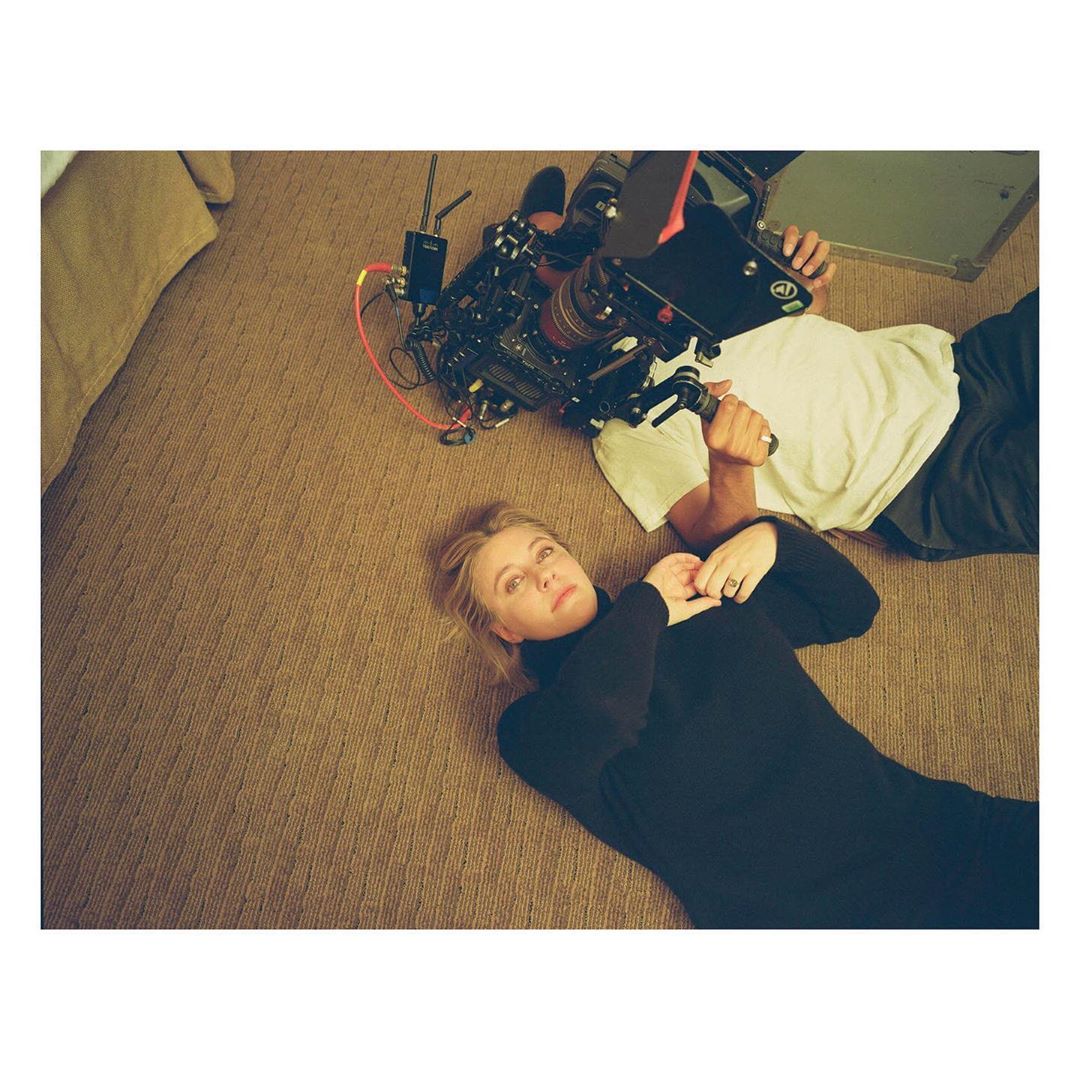 Adult woman lying on a patterned carpet, looking up at a camera operator with equipment, creating behind-the-scenes vibe in an indoor setting.