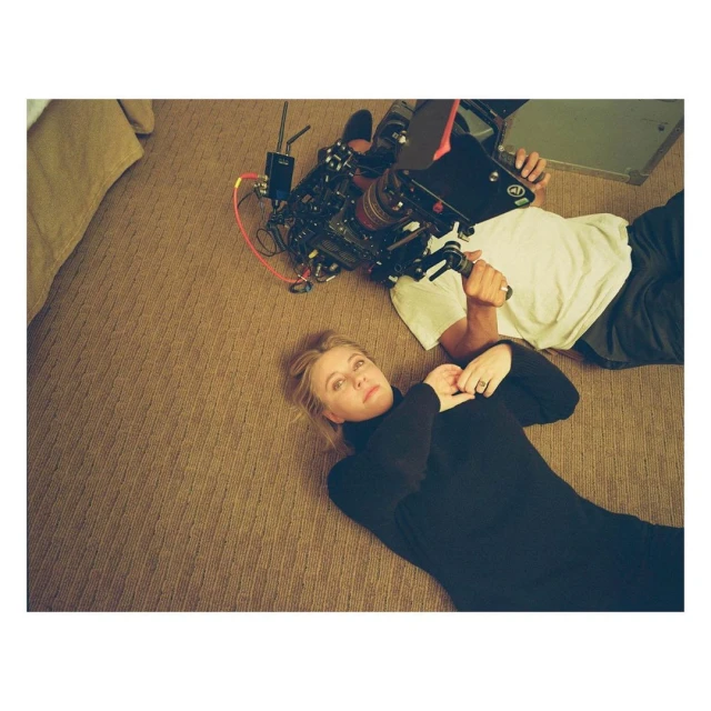Adult woman lying on a patterned carpet, looking up at a camera operator with equipment, creating behind-the-scenes vibe in an indoor setting.