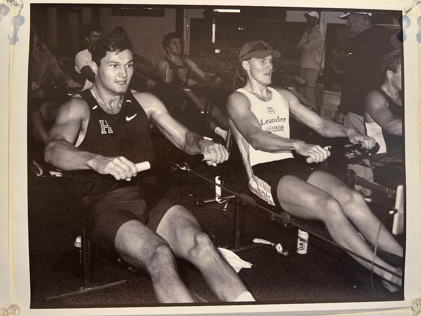 Black and white photo of two adult males using rowing machines in an indoor gym setting with other people visible in the background.