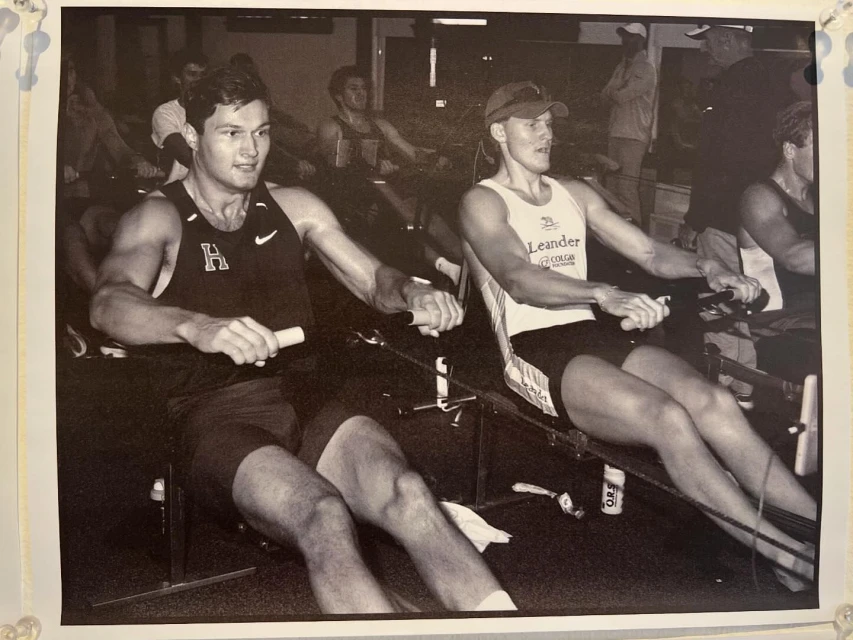 Black and white photo of two adult males using rowing machines in an indoor gym setting with other people visible in the background.