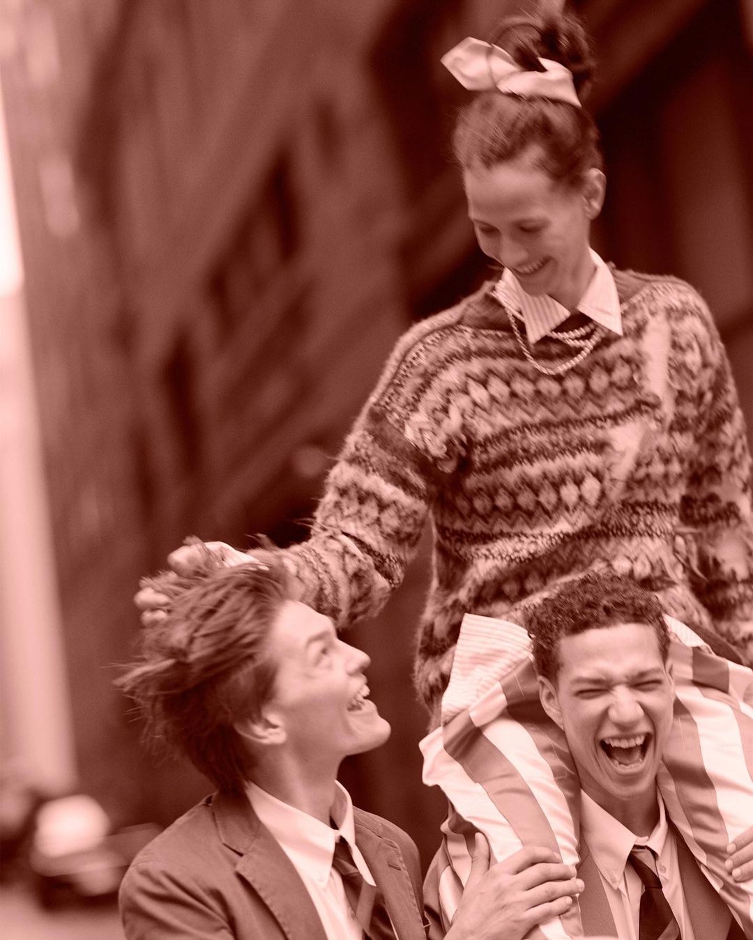 Three smiling people pose outdoors in a cheerful group shot. One woman stands and two men, wearing suits, are positioned below her.