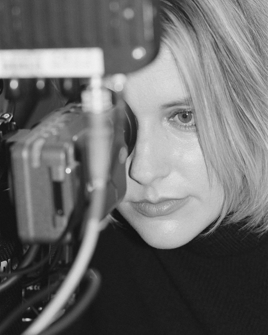 Monochrome portrait of a blonde woman in a black top looking through a professional film camera, with visible camera equipment.
