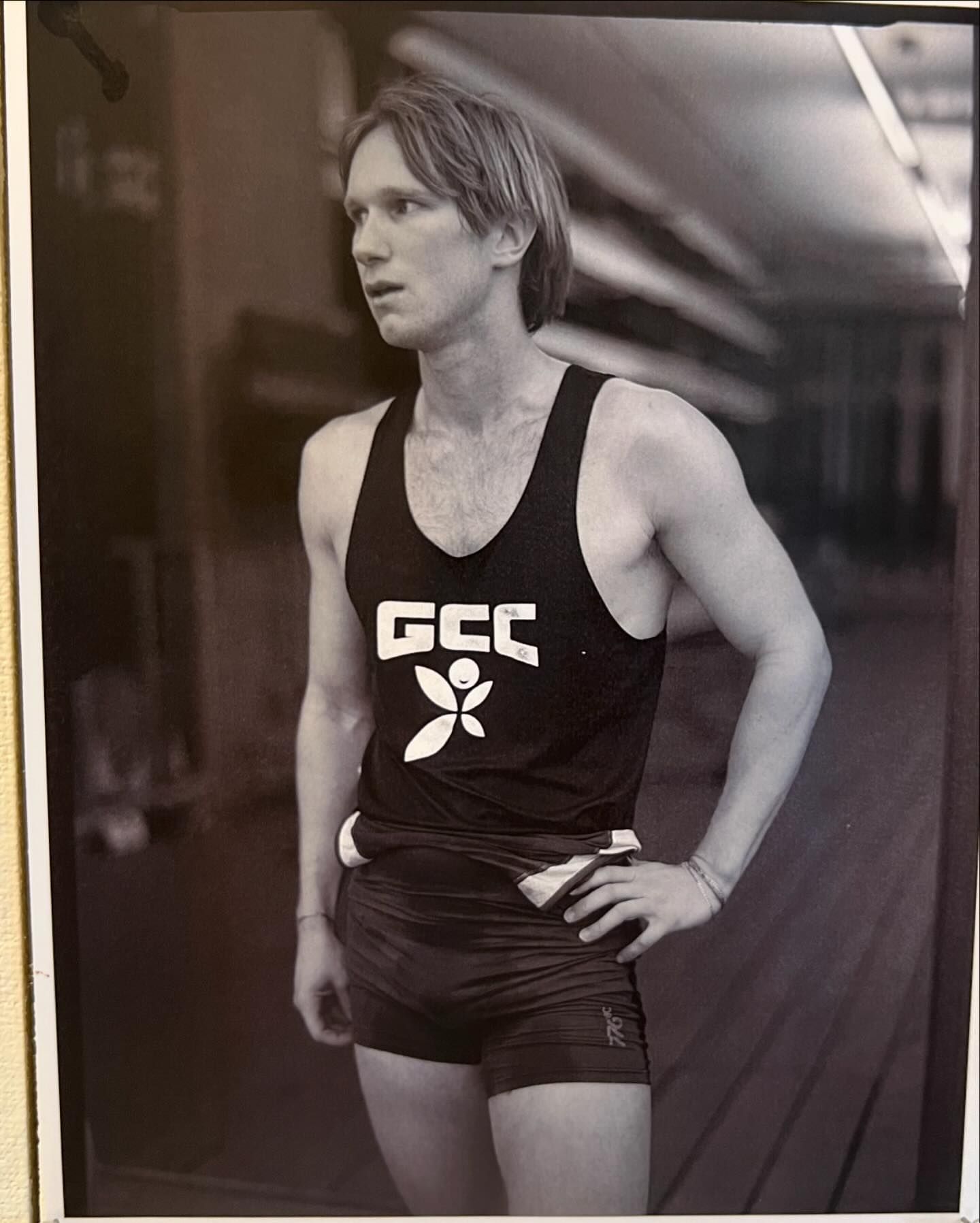 Black and white photo of a blond male athlete in a dark singlet and shorts, gazing left with his hand on his hip.