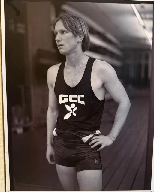 Black and white photo of a blond male athlete in a dark singlet and shorts, gazing left with his hand on his hip.